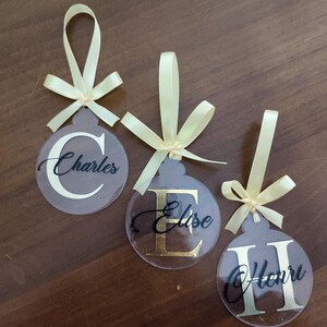 以下が含まれることがあります： 金色と黒色の文字が書かれた透明なプラスチック製のクリスマスオーナメントが3つ。オーナメントは黄色のリボンで飾られています。オーナメントには「チャールズ」、「エリーゼ」、「ヘンリー」という名前が書かれています。