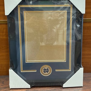May include: A black picture frame with a gold mat and a gold Notre Dame Fighting Irish logo. The frame is wrapped in plastic.