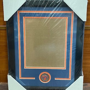 May include: A black picture frame with a blue mat and orange trim. The frame has a clear plastic cover and a small New York Knicks logo in the bottom center.