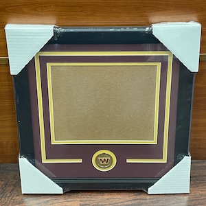 May include: A framed picture with a black border, maroon matting, and gold accents. The frame has a team logo at the bottom. The frame is wrapped in protective packaging.