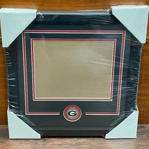 May include: A black picture frame with a red and white border. The frame has a gold circular emblem with a white letter "G" in the center. The frame is wrapped in plastic.