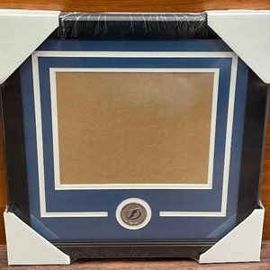 May include: Black framed picture frame with a blue and white matting. The frame has a silver emblem at the bottom center. The frame is wrapped in plastic and protected by white corner protectors.