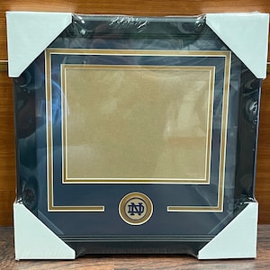 May include: A black picture frame with a gold trim and a gold Notre Dame logo in the bottom center. The frame is in a protective plastic wrapping.