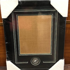 May include: A black picture frame with a gold trim and a silver Philadelphia Eagles logo in the center. The frame is made of wood and has a glass front.