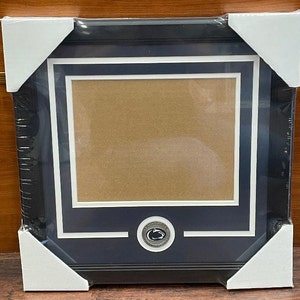May include: A framed picture with a navy blue border, white matting, and a brown interior. The frame has a silver emblem at the bottom center. The frame is wrapped in clear plastic and has white corner protectors.