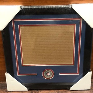 May include: A black picture frame with a blue and red mat. The frame has a small, round, silver-colored Chicago Cubs logo in the bottom center.