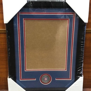 May include: A black framed picture with a blue mat and red trim. The mat has a circular cutout with a silver Chicago Cubs logo. The picture is ready for a photo or other memorabilia.