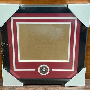 May include: A black framed picture with a red mat and a white inner mat. The picture has a gold medallion with a baseball logo in the center.