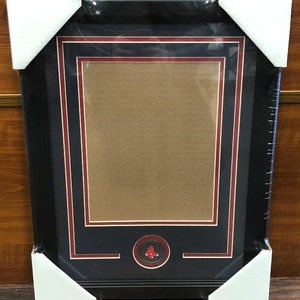 May include: A black picture frame with a red and white border. The frame has a small, round, red and white emblem in the bottom center. The emblem has a red baseball logo.