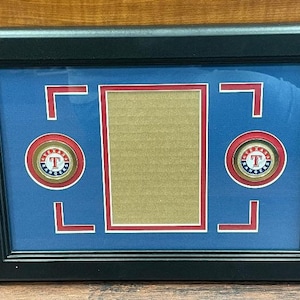 May include: A black framed picture with a blue mat and two gold and red baseball team logos. The logos are on a white background with the letter "T" in the center. The picture has a blank space in the center.