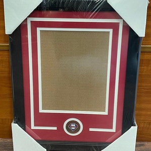 May include: A black picture frame with a red mat and white border. The frame has a circular emblem with the words "Philadelphia Phillies" in the center.
