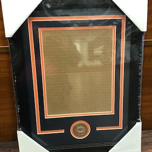 May include: A black picture frame with a double orange and black mat. The frame has a small, round, bronze-colored medallion with the Chicago Bears logo in the center.