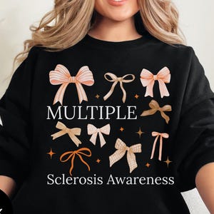 May include: Black sweatshirt featuring a design of various bows in shades of peach, tan, and brown. The words "MULTIPLE Sclerosis Awareness" are printed in white. The sweatshirt has a round neckline.