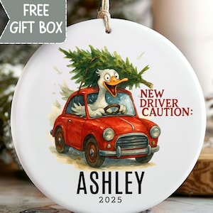 May include: White ceramic Christmas ornament with a cartoon duck driving a red car with a Christmas tree on top. Text reads "NEW DRIVER CAUTION:" and "ASHLEY 2025". Includes a "FREE GIFT BOX" graphic.