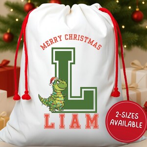 May include: White Christmas gift bag with red drawstring, featuring a green letter "L" and a cartoon dinosaur wearing a Santa hat and Christmas lights. The text "MERRY CHRISTMAS" and "LIAM" are in red.