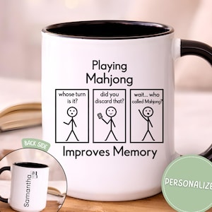 May include: White ceramic mug with a black handle and rim, featuring the text "Playing Mahjong Improves Memory." The design includes stick figures and phrases like "whose turn is it?" and "did you discard that?" A smaller image shows the back with a name.