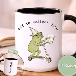 May include: White ceramic mug with a black interior and handle. The mug features a whimsical illustration of a frog riding a scooter, wearing a wizard hat and glasses, and holding a chart. Text above the frog reads "off to collect data."