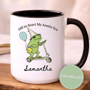 May include: White ceramic mug with a black handle and rim. The mug features a green frog riding a scooter, wearing a hat and holding a balloon. Text on the mug reads "Off to Start My Auntie Era" and "Samantha". A green circle says "Personalized".