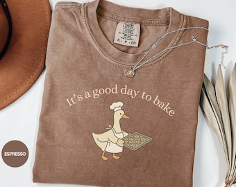 May include: Espresso-colored t-shirt with the phrase "It's a good day to bake" and a cartoon duck wearing a chef's hat and apron, holding a tray of cookies. A silver necklace with a pendant is on the shirt.