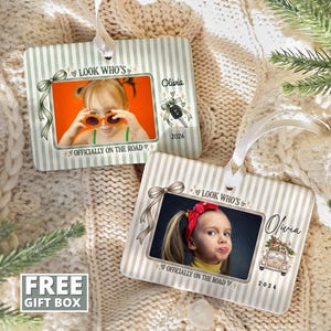 May include: Two rectangular ornaments with white ribbon hangers, featuring a light green and white striped background. Each ornament displays a photo of a child, the text "LOOK WHO'S OFFICIALLY ON THE ROAD", the name "Olivia", the year "2026", and a car key or car illustration.
