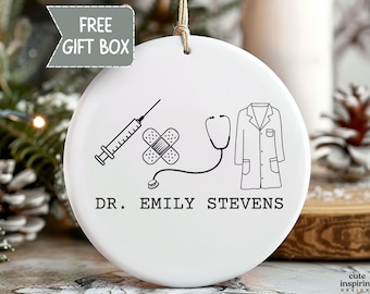 May include: White ceramic ornament with a medical theme. Features a syringe, bandage, stethoscope, and lab coat illustrations. The text "DR. EMILY STEVENS" is printed below the illustrations. Includes a "FREE GIFT BOX" tag.