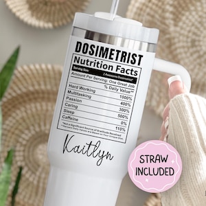 May include: White tumbler with a handle and a straw, featuring the text "DOSIMETRIST Nutrition Facts." The label lists daily values for hard work, multitasking, passion, caring, sleep, and caffeine. A pink sticker says "STRAW INCLUDED."