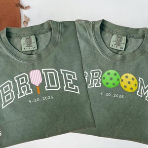 May include: Two moss green t-shirts with white text and graphics. One shirt says "BRIDE" with a pink and orange pickleball paddle. The other says "GROOM" with green and yellow pickleballs. Both shirts have the date "4.20.2026".