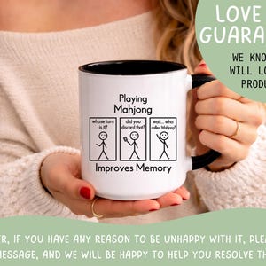 May include: White ceramic mug with a black interior and handle. The mug features the text "Playing Mahjong Improves Memory" with stick figure illustrations and the phrases "whose turn is it?", "did you discard that?", and "wait... who called Mahjong?"