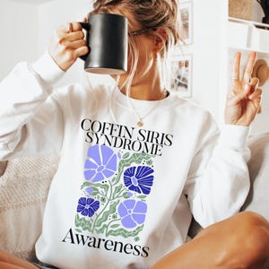 May include: White sweatshirt with the text "COFFIN SIRIS SYNDROME" above a floral design in blue, purple, and green. The word "Awareness" is printed below the floral design. A black mug is held up near the face.