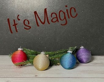 Könnte beinhalten: Fünf Weihnachtskugeln in einer Reihe auf einer weißen Holzoberfläche. Die Kugeln sind rot, gold, blau, lila und grün. Die Kugeln sind alle rund und haben eine glänzende Oberfläche. Der Text "It's Magic" ist auf einer schwarzen Tafel im Hintergrund geschrieben.