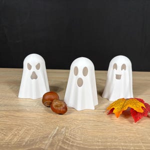 K&ouml;nnte beinhalten: Drei wei&szlig;e Geisterfiguren mit ausgeschnittenen Gesichtsz&uuml;gen. Der linke Geist hat einen w&uuml;tenden Ausdruck, der mittlere Geist hat einen ovalen Mund und der rechte Geist hat ein l&auml;chelndes Gesicht. Zwei braune Kastanien und ein kleines orange-rotes Blatt.