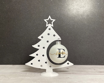 Könnte beinhalten: Weißer Weihnachtsbaum-Schmuckhalter aus Metall mit einem weißen Ornament mit dem Buchstaben E und dem Namen "Emmeline" in goldener Schrift.