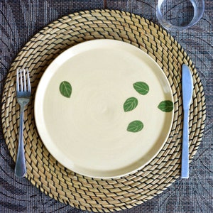 May include: A cream-colored ceramic plate with green leaf designs, set on a woven placemat. A silver fork and knife are placed on either side of the plate. The placemat rests on a patterned tablecloth.