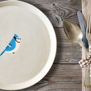 Könnte beinhalten: Ein weißer Keramikteller mit einer Illustration eines Blauhähers. Der Teller steht auf einem Holztisch mit einer braunen Leinenserviette und silbernem Besteck.