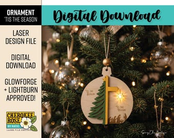 May include: A round wooden Christmas ornament featuring a layered green pine tree, a yellow cross, and a star cutout. The text 'Tis the season' and 'Tis the reason' is engraved alongside a silhouette of a nativity scene at the bottom.