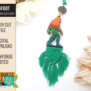 May include: A laser cut wooden Bigfoot charm with a green and orange gradient, hanging from a green macrame tassel. The charm is labeled "BIGFOOT CAR CHARM/DIFFUSER" and "LASER CUT FILE".
