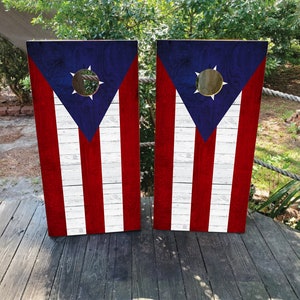 May include: Two wooden cornhole boards with a distressed white and red woodgrain background. The boards are decorated with the Puerto Rican flag, featuring a blue triangle with a white star in the centre.