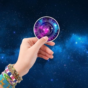 May include: A hand holding a round sticker with a galaxy design. The sticker has a white border and a pink, blue, and purple galaxy design with white lines.