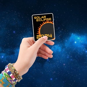 May include: A hand holds up a black card with the words "Solar Eclipse 2024" and a graphic of a sun with a black circle in the center.