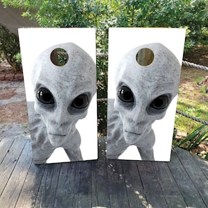 May include: Two white cornhole boards with a close-up image of a gray alien with large black eyes. The alien is looking directly at the viewer.
