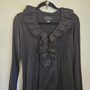 May include: A black long-sleeve shirt featuring a deep V-neckline adorned with a prominent, layered fabric ruffle. The garment includes a label at the inner neckline with the text Lauren Jeans Co. and long sleeves with small buttons at the cuffs.