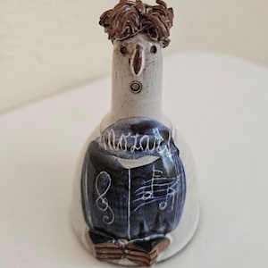 May include: A ceramic figurine of a figure with brown hair, a surprised expression, and a blue and white outfit. The figurine has the word "Mozart" written on the front, along with musical notes.