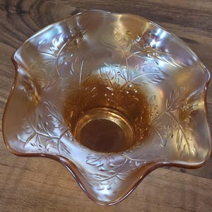Op de afbeelding: Een iriserende oranje glazen kom met een gegolfde rand. De kom heeft een gedetailleerd blad- en bessenpatroon. De binnenkant van de kom is donkerder oranje en de buitenkant heeft een glinsterende, reflecterende kwaliteit. De kom is waarschijnlijk vintage.