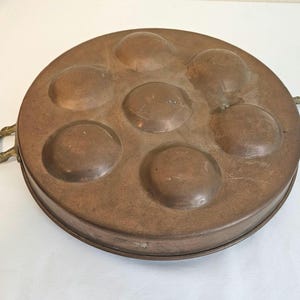 Puede incluir: Una sartén redonda de cobre para caracoles con siete compartimentos empotrados. La sartén tiene dos asas de latón y un borde ligeramente elevado. El cobre tiene una pátina cálida y envejecida, lo que sugiere que es vintage o antiguo.