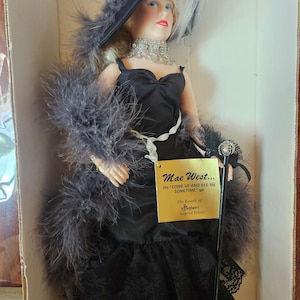 May include: A Mae West doll in a box, dressed in a black gown with a feathered boa and hat. The doll has blonde hair, red lipstick, and a jeweled necklace. A tag reads "Mae West... the 'COME UP AND SEE ME SOMETIME' gal."