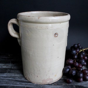 以下が含まれることがあります： 白い陶器製の壺で、持ち手は一つです。壺は使い込まれたような、素朴な風合いがあり、暗い木製の表面の上に置かれています。壺の隣には、濃い紫色のぶどうの房が置かれています。