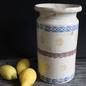 May include: A white ceramic jar with blue and brown geometric patterns and yellow accents. The jar has a wide mouth and a slightly rounded body. Three lemons are in the foreground.