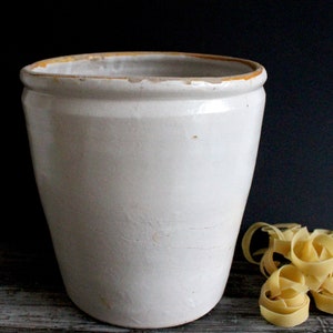 Op de afbeelding: Een witte keramische pot met een licht verweerde afwerking. De pot heeft een brede opening en een afgeronde basis. Een kleine hoop gele pasta ligt naast de pot, wat wijst op een keuken- of eethema.