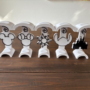 May include: Five white plastic stands featuring different Disney character silhouettes. The characters are Mickey Mouse, Minnie Mouse, Rapunzel, Ariel, and Cinderella's castle.