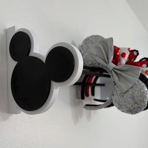 May include: A white wall-mounted organizer shaped like Mickey Mouse's head, with black ears. It holds several decorative headbands, including a silver sequined pair with a large bow, and a red and white polka-dot pair.